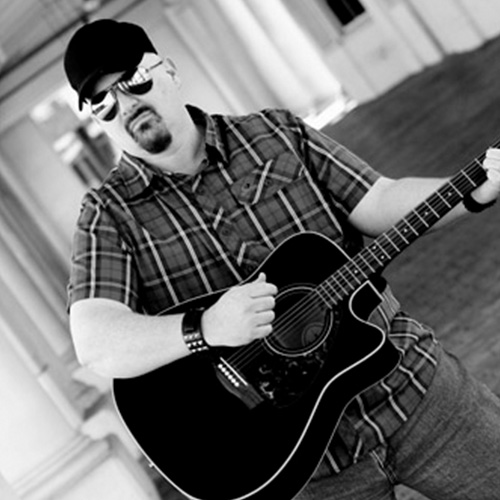 A portrait of Thad Foster in a baseball cap and aviator glasses holding a guitar.