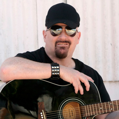 A portrait of Thad Foster in a baseball cap and aviator glasses holding a guitar.
