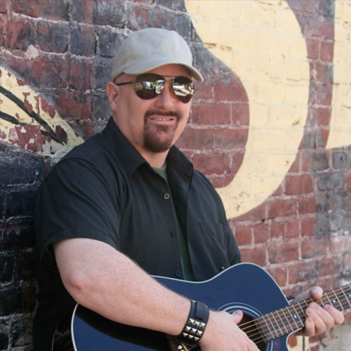 A portrait of Thad Foster in a baseball cap and aviator glasses holding a guitar and standing against a brick wall.