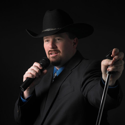 A portrait of Thad Foster in a hat holding a microphone in one hand and a microphone stand in the other hand and singing.